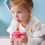 Petite fille rosse qui boit dans un verre à l'aide d'une paille orange et d'un couvercle sur le verre rose