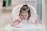 Petite file assise sur sa chaise haute, entrain de manger du yaourt dans un bol blanc avec des couverts de couleur rose.