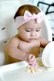 Bébé assit sur une chaise haute entrain de manger des pates avec une fourchette de couleur rose. La petite fille a un bandeau de couleur rose