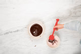 Photo prise par le dessus d'une table de cuisine en marbre avec posé dessus un bol avec du chocolat fondut dedans. Il y a une main d'enfant qui tient une spatule de cuisine de cuoleur rouge qui reverse le chocolat dans un autre moule