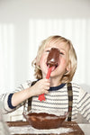 Petit garçon blond qui lèche une cuillère de cuisine rouge, pleine de chocolat fondut. Devant lui est posé sur une table un petit saladier avec du chocolat à l'intérieur