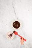 Photo prise par le dessus d'une table de cuisine en marbre avec posé dessus un bol avec du chocolat fondut dedans. Il y a une main d'enfant qui tient une spatule de cuisine de cuoleur rouge qui reverse le chocolat dans un autre moule
