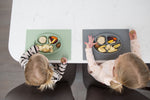 Photo prise au dessus d'une table ou 2 enfants mangent avec leurs main dans un plateau en silisone avec assiettes anti dérapantes
