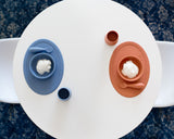 Photo prise de haut d'une table oval avec posé dessus à chaque extrimité de la vaisselle identique mais de 2 couleurs differentes : rose et bleu. il y a un set de table avec bol intégré, une cuillère et un gobelet