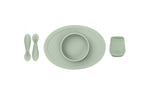 Photo sur fond blanc d'un set de repas composé d'une assiette avec set de table et bol integré, ainsi que de 2 petites cuillères et une petite tasse. Le set est de couleur vert amande et c'est en silicone