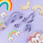 Photo d'ustensiles de cuisine posé sur un fond violet, il y a des emportes pièces an acier en forme de puzzle pour réaliser une licorne et un arc en ciel en 3D. A coté sont posés les biscuits réalisés et décoré avec de la pâte à sucres