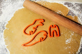 Plan de travail cuisine avec pâte à  sablées étalées avec rouleau a pâtisserie. Sur la pâte a sablées il y a 3 emportes-pièces rouge en forme de dinosaure.