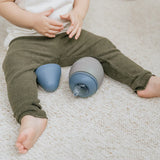 photo d'une gourde pour enfant posée sur un tapis blanc, la gourde est entre les jambes d'un bébé, on apperçoit uniquement ses jambes. La gourde est de forme arrondie, elle est grise avec un couvercle de couleur bleu