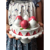 Photo d'un jouet en bois pour enfant en forme de gâteau aux fraises tenu dans les main d'une petite fille. L'angle de la photo est prise sur les mains et le gâteau, on ne distingue pas le visage de la jeune fille