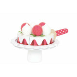 Jouet pour enfant en bois en forme de gâteau aux fraise. Le gâteau est posé sur un plateau de présentation blanc, il se coupe en 6 part et il dispose de fraise et de meringue sur le dessus. Devant le gâteau est posée une pelle à gâteau