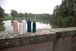 Photo de 5 gourdes de 3 couelurs différentes et tailles differentes (rose, blanche et blleu) posées sur un muret en pierre avec en arrière plan un paysage de campagne avec un étang et des arbres