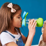 Photo d'une jeune fille de profil qui est entrain de boire dans une gourde en plastique auc couleurs vert et bleu. La fille est coifée avec un petit noed rose dans les cheveux