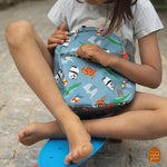 Enfant sur un skate-board bleu assit en tailleur. L'enfant porte sur ses genoux un sac a dos bleupétrol avec des animaux dessus.