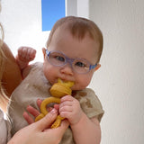 Photo d'un bébé qui porte des lunettes violettes et qui porte a sa bouche avec l'aide de sa maman une grognoteuse de couleur jaune