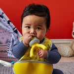 Bébé habillé d'un pull gris avec un bavoir jaune autour du cou. Le bébé porte a sa bouche une sucette à aliment de couleur assorti au bavoir