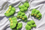 7 jouets pour bébé en forme d'endamame posés sur une nape de couleur blanche. les jouets sont en caotchouc et ressemble parfaitement au vrai légumes