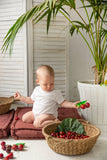Photo d'un bébé habillé en body blanc qui tient dans sa main un anneau de dentition en forme de cerise et qui a autour de lui un panier en osier replit de cerise