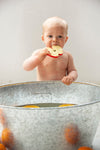 Photo d'un bébé torse nu derrière une grosse bassine en inox, le bébé porte a sa bouche un jouet de dentition en forme de tranche de pomme