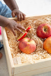 Photo d'un panier en bois remplit de pommes avec de la paille, dans le panier il y a une tranche de pomme coupé qui est un jouet de dentition pour bébé. La tranche de pomme est saisit par la main d'un bébé