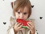 Phot d'une petite fille devant un papier peint avce des petit coeur, l'enfant porte à sa bouche un jouet en forme de tranche de pastèque