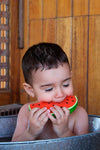 Photo d'un jeune garçon assit dans une bassine en inox, le garçon ferme les yeux et porte à sa bouche un jouet en caoutchouc en forme de pastèque