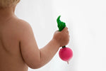 Photo d'un bébé de dos qui tient dans sa main un radis en caoutchouc, le jouet ressemble trés fortement à un vrai legume. La photo est centré sur le bras du bébé, on ne distingue pas le visage de l'enfant
