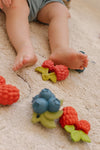 Phot d'un tapi avec un bébé dessus, on aperçoit uniqument ses jambes et pieds. Sur le tapis sont posés des jouets en forme de fruits : myrtilles et framboises