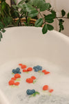 Photo d'une baignoire avec de l'au et de la mousse. Dans l'eau il ya des jouets de bain en forme de framboises et de myrtilles. Derrière la baignoire il y a une plante verte
