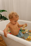 Photo d'un bébé dans un bain avec dans l'eau de son bain, tous plein de jouets de bain en forme de fruits et légumes. Le bébé est un garçon et il porte un maillot de bain short bleu 