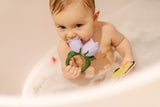 Bébé dans un bain avec de la mousse, il tient à sa main un anneau de dentition en forme de fleur blanche et un papillon jaune dans l'autre. La fleur represente une campanule