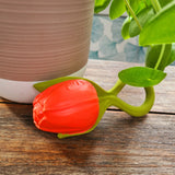Photo de jouet de dentition représentant à la quasi perfection des fleurs, il y a 2 fleurs mauve et une tulipe rouge. Les fleurs sont posées sur une table en bois avec une plante verte en arrière plan