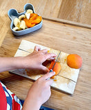Main d'enfant qui coupe un abricot avec un couteau pour enfant de couleur gris. Une petite boite en silicone est sur le haut gauche de la photo, ou il y a le fruits coupé