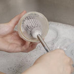 Gros plan sur les main d'un adulte qui nettoit un recypient dans un lavabo remplit de mousse, à l'aide d'un goupillon avec une tête en silicone