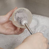 Gros plan sur les main d'un adulte qui nettoit un recypient dans un lavabo remplit de mousse, à l'aide d'un goupillon avec une tête en silicone