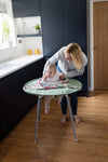 Photo d'une maman qui instale un bébé sur une chaise haute équipé d'un plateau repas sauge avec un tablier. La photo est prise dans une cuisine avec des porte bleu marine