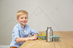 Photo d'un garçon souriant (avec 2 dents en moins) assis à une table avec posé devant lui une lunch box ainsi qu'une gourde en inox ouverte avec son bouchon posé a coté. Le garçon regarde l'objectif, il porte une chemise bleu avec un colier