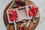 Photo d'une planche en bois de cuisine avec posé dessue un moule à glace rose contenant 2 glaces à l'eau et aux morceaux de fraises. Les glaces sont en forme d'animaux. Des fraises entières sont également posés sur la planche