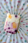 Photo d'une glace au yaourt en forme de tête de panda. ELle contient des morceaux de fruits jaune et violet. En arriere plan on voit flouté un éventail blanc avec des pois de couleurs rose, vert et jaune