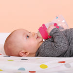 Bébé allongé sur un tissu blanc à pois de couleur entrain de boire au biberon seul en tenant les poignée du biberon