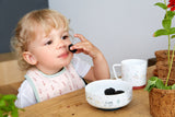 Jeune enfant blond assit a une table qui porte une mur a sa bouche. Devant l'enfant est posé de la vaisselle en porcelaine de couleur blanche avec des motifs nature. 