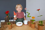 Photo d'une jeune fille souriante blonde, qui tient dans sa main une brochette avec un cartier de pastèque. Elle se trouve devant une table ou est posé desss plusieurs pot contenant une fleurs chacun. il y a egalement un bol ainsi qu'une tasse devant elle