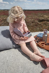Photo d'une jeune fille blonde habillé d'une rose rose et coiffée vec un bandeau rose. Elle est assise sur un coussin par terre dans un champ, elle tient sur ses genoux une boite à gouter, à coté d'elle il y a un plateau avec un pic nique posé dessus