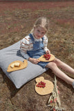 lp Photo d'une jeune fille assise sur un gros coussin dans un champ avec devant elle et sur ses genoux des planches en bois contenant du raisin et de la brioche. La jeune fille est souriante et vêtue d'une salopette en jeans
