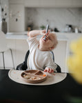 Jeune garçon assit à table et entrain de manger. il a devant lui une assiette compartimenté contenant de la nourriture. il tient dans sa bouche une cuillère et leve la tête en fermant les yeux