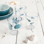 Photo de 3 verres pour enfant, les verres sont transparent avec des illustration d'animaux marin bleus. Les verres sont sur une table en bois blanche avec posé dessus des coquillages blancs et de la vaisselle bleu