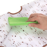 Gros plan sur la main d'un adulte qui tient une lavette verte dans la main et qui semble nettoyer un tissus blanc avec des illustration d'oies dessus