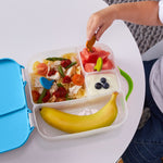 Photo d'une lunch box d'enfant avc la main d'un enfant qui mange à l'aide d'une mini fourchette. La lunch box est compartimentée, elle contient de la salade de pates, du yaourt, une banane et des morceaux de pastèques