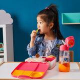 Jeune fille assise à table qui mange dans une lunch box compartimentée, elle a devant elle une gourde qui contient de l'eau et qui a les même couleurs que sa boite à repas