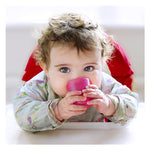 Photo d'un bébé qui est assit sur une chaise haute, il regarde l'objectif. il a les yeux clair, il est habillé d'un chemisier avec des motifs fleuris. Il est entrain de boire dans une tasse de couleur rose