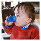 Photo d'un bébé qui boit dans un petite verre bleu. Le garçon a un tablier de couleur rouge avec les manche et le col bleu